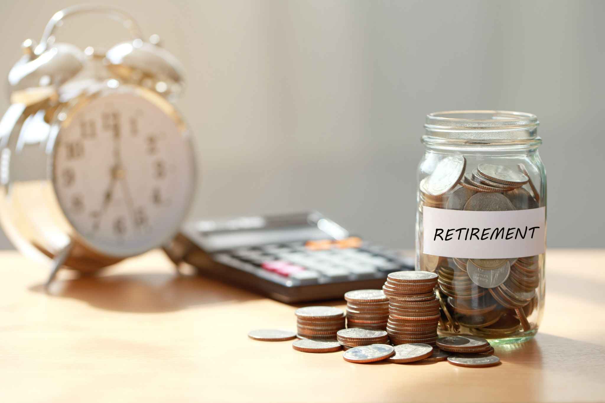 צנצנת זכוכית מלאה במטבעות עם תווית "RETIREMENT", שעון מעורר מטושטש ומחשבון ברקע – קונספט של חיסכון לפנסיה ותכנון פרישה.